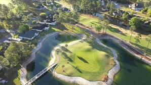 An aerial view of a golf course with a pond, surrounded by residential houses and a bridge.