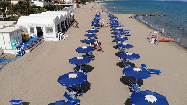 A beach with rows of blue umbrellas and chairs, a white building with a terrace, and a view of the sea and mountains in the background.