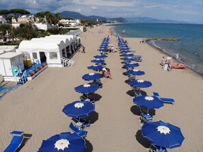 A beach with rows of blue umbrellas and chairs, a white building with a terrace, and a view of the sea and mountains in the background.