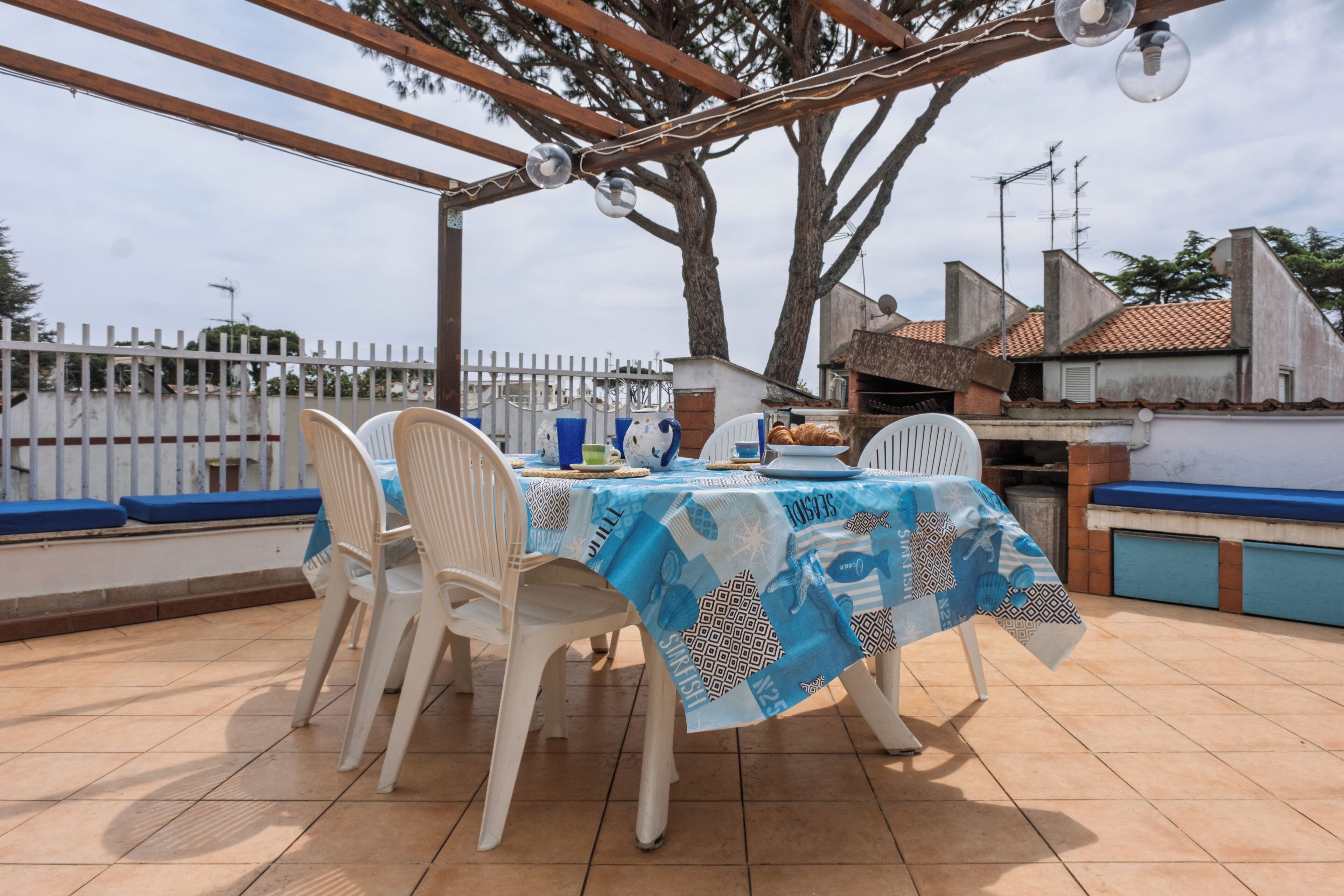 Une terrasse extérieure avec une table recouverte d’une nappe bleue, entourée de chaises en plastique blanches.