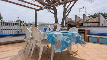 Une terrasse extérieure avec une table recouverte d’une nappe bleue, entourée de chaises en plastique blanches.
