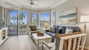 A spacious living room with a balcony, a ceiling fan, a television, and a painting of a beach scene.
