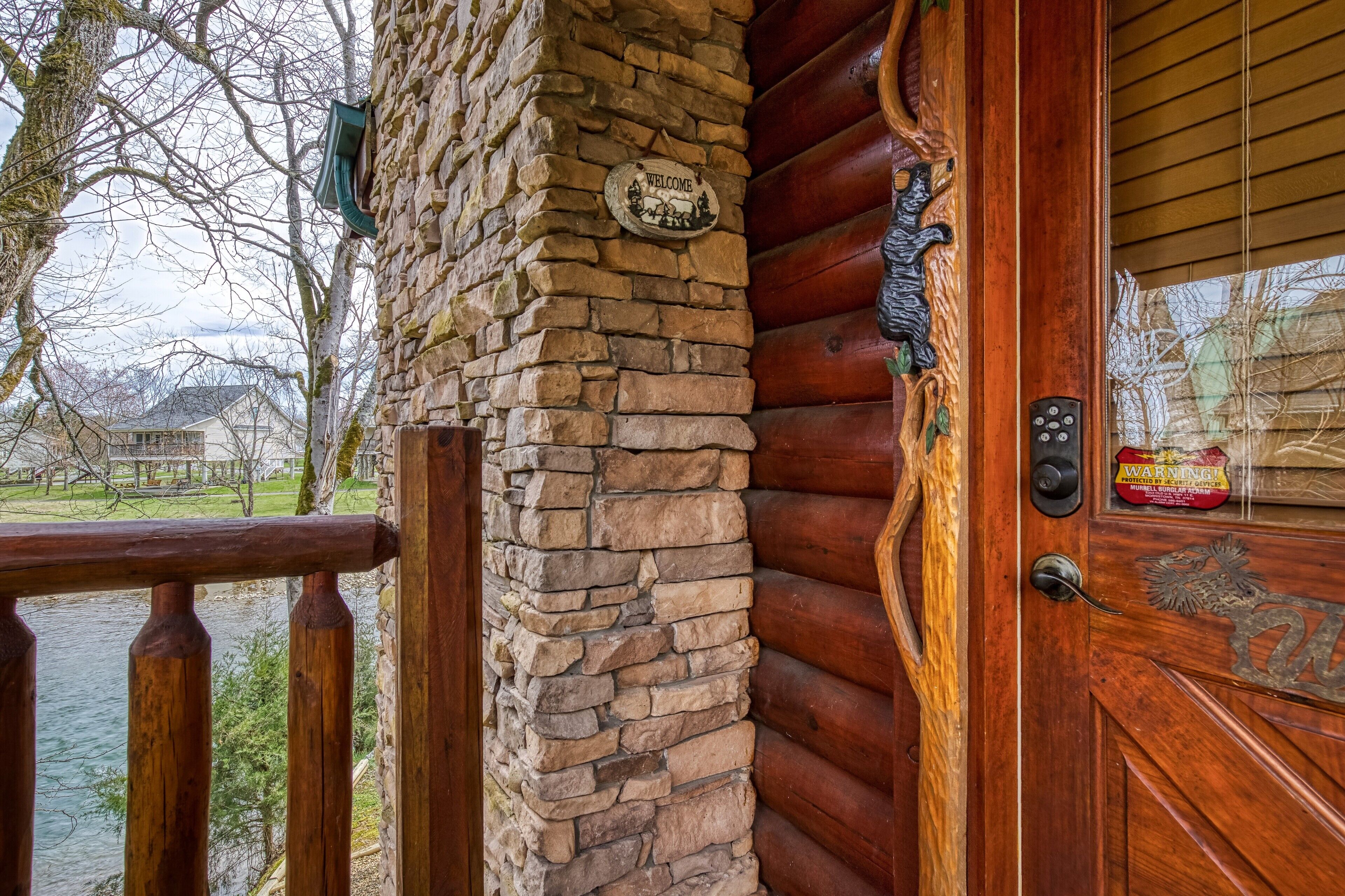 Une cabane en rondins avec un mur en pierre, une porte en bois ornée d’un panneau d’avertissement et une vue sur un lac.