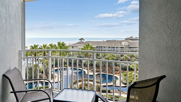 Un balcon donnant sur une piscine et des palmiers, avec vue sur un immeuble doté d’un toit plat.