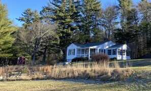 A single-story house with a porch, surrounded by trees and a grassy field.