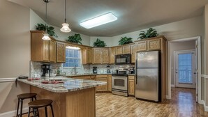 A kitchen with wooden cabinets, a central island with stools, stainless steel appliances, and a bright ceiling light. - Pigeon Forge Cottage w/ Hot Tub & City Lights, Near Dollywood Sleeps 7 (PIGEON FORGE, TN)