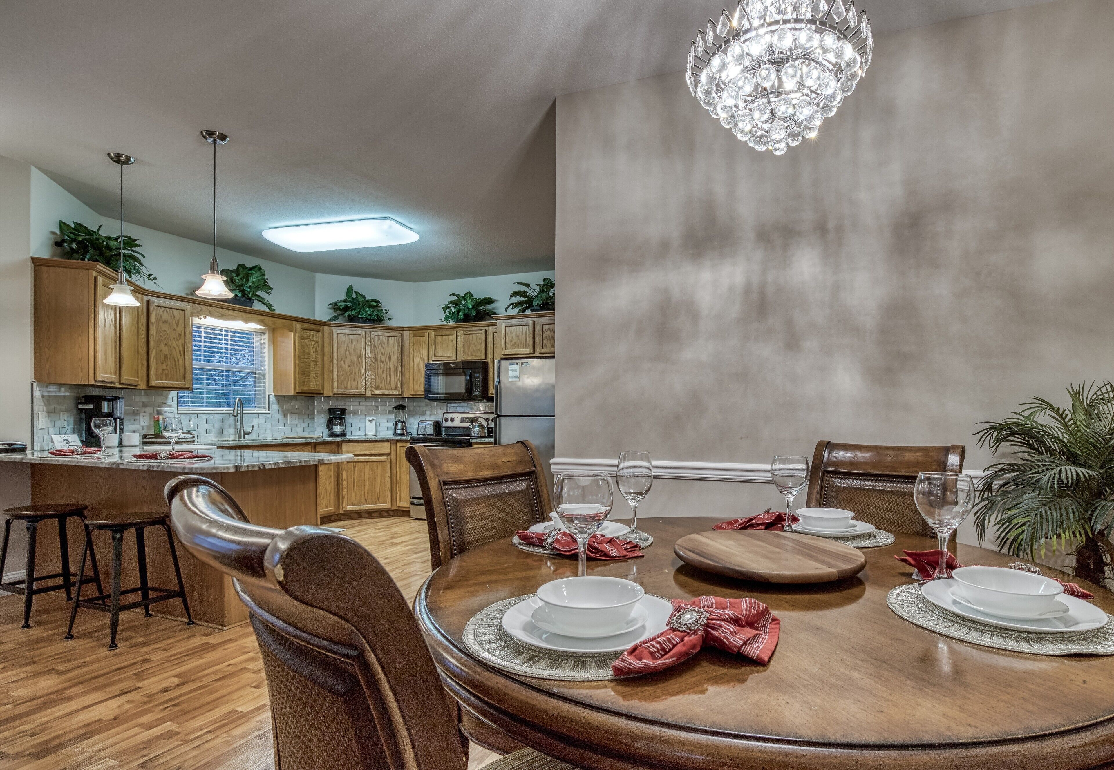 A dining area with a wooden table set for four, surrounded by chairs. A chandelier hangs above the table, and the room has wooden cabinets and a window with blinds.