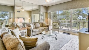 Ein geräumiges Wohnzimmer mit einem großen Glasfenster, das auf einen Außenbalkon führt, mit Sofas, einem Couchtisch und einem Fernseher möbliert.