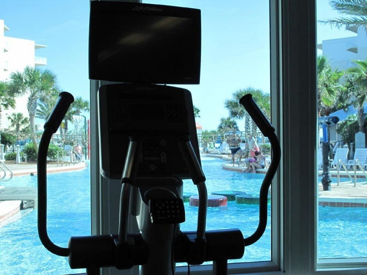 Una bicicleta estática con panel de control, ubicada cerca de una ventana con vista a una piscina con sillas de descanso y palmeras.