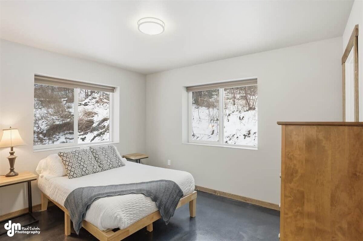 A bedroom with a bed, two windows with snowy views, a nightstand, a lamp, and a wooden dresser.