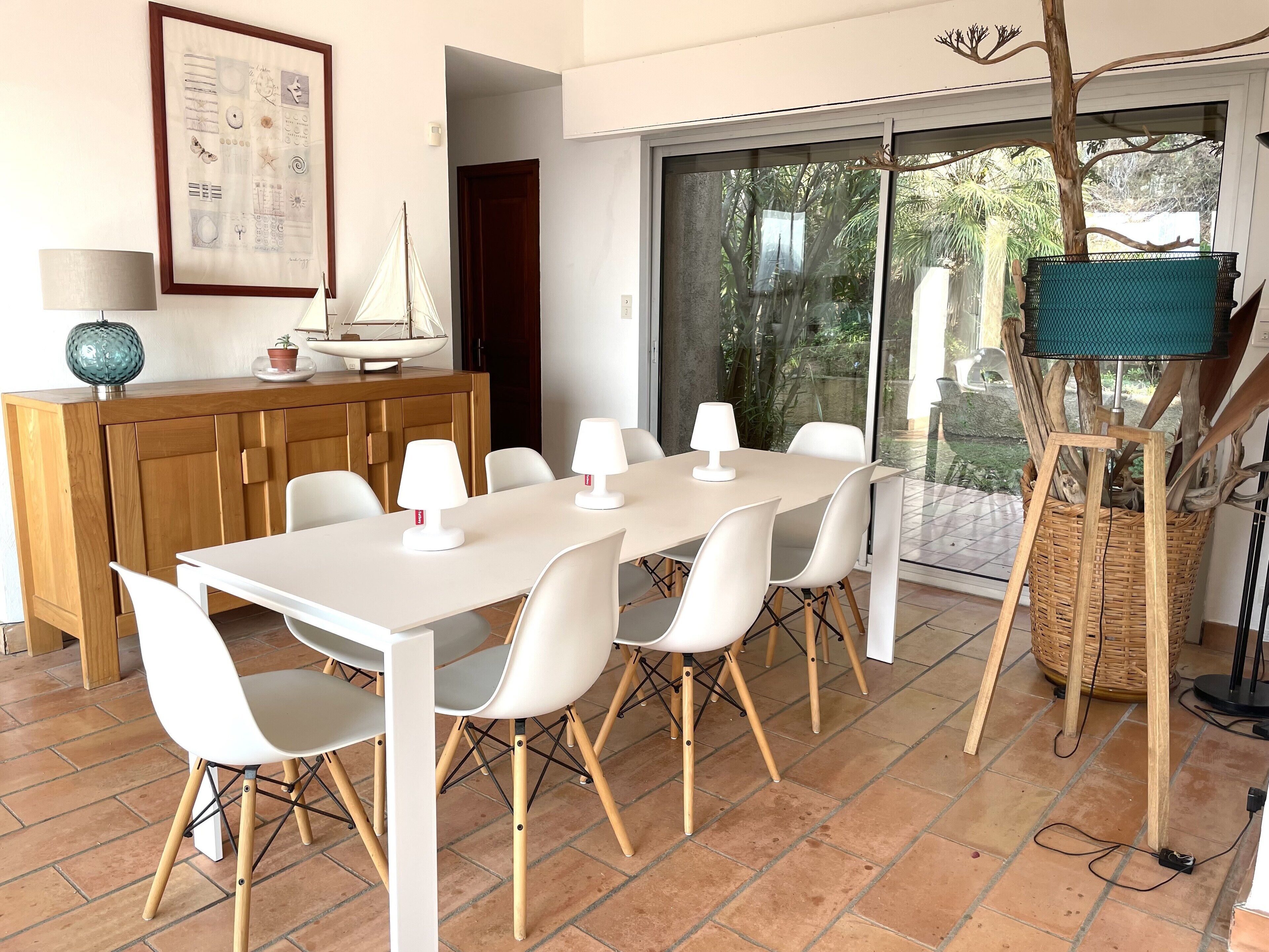 A dining area with a white table and chairs, a wooden cabinet, a framed artwork, and a view through a glass door.