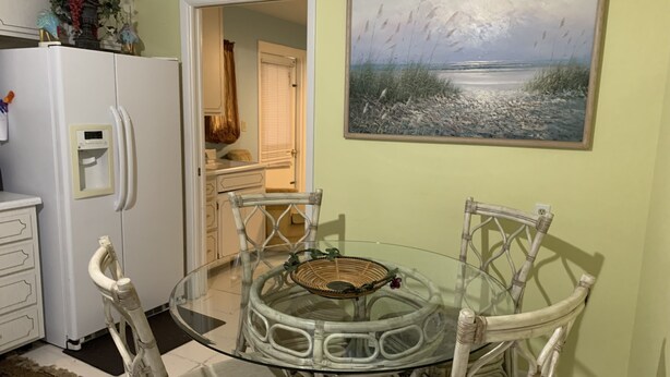 A dining area with a glass table surrounded by wicker chairs, a white refrigerator, and a painting of a beach scene. - Clean And Comfortable  (Sebring)