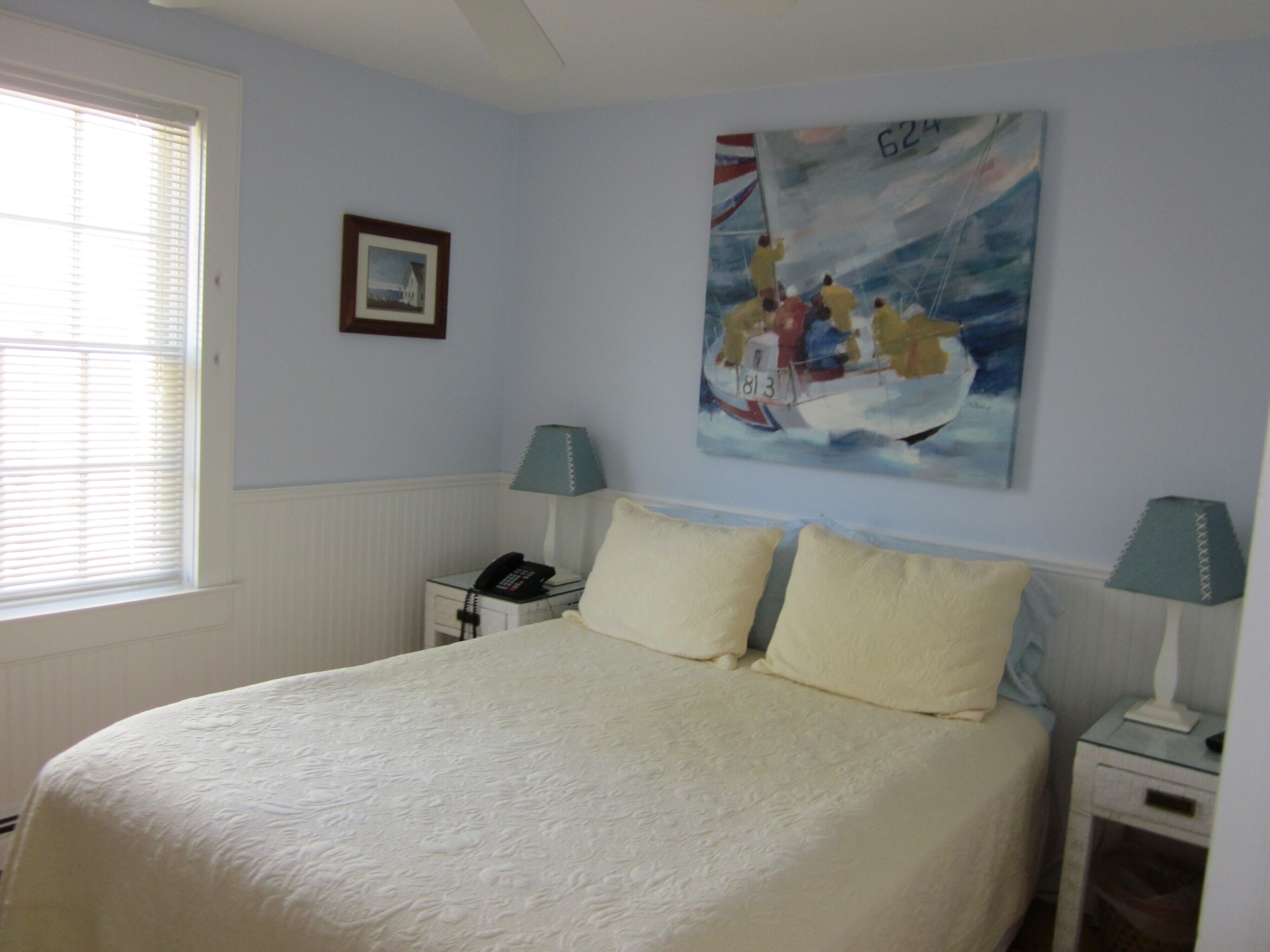 A bedroom with a bed, two bedside tables, a nightstand, a window with blinds, and a painting on the wall.