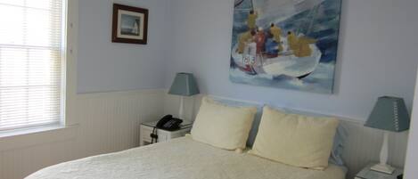 A bedroom with a bed, two bedside tables, a nightstand, a window with blinds, and a painting on the wall.