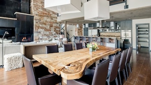 Un vaste espace dédié à la salle à manger, avec une grande table en bois et des chaises modernes, une cheminée en pierre et une cuisine en arrière-plan.