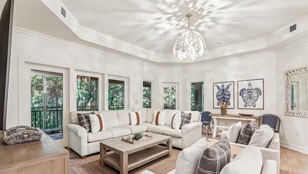 A spacious living room with a large sectional sofa, a coffee table, and a chandelier. There are multiple windows providing natural light and views of greenery outside. - Picturesque Villa w/ Screened Porch & Amenities (Hilton Head Island)