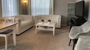 A living room with two beige sofas, a white coffee table with plants, a wooden table, and a television on a stand. - 4 bedroom townhouse at The Villas at seven Dwarfs (Kissimmee)