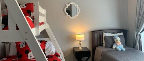 A child's bunk bed with Mickey Mouse bedding, a bedside table with a lamp, a mirror on the wall, and a bed with a plush toy.