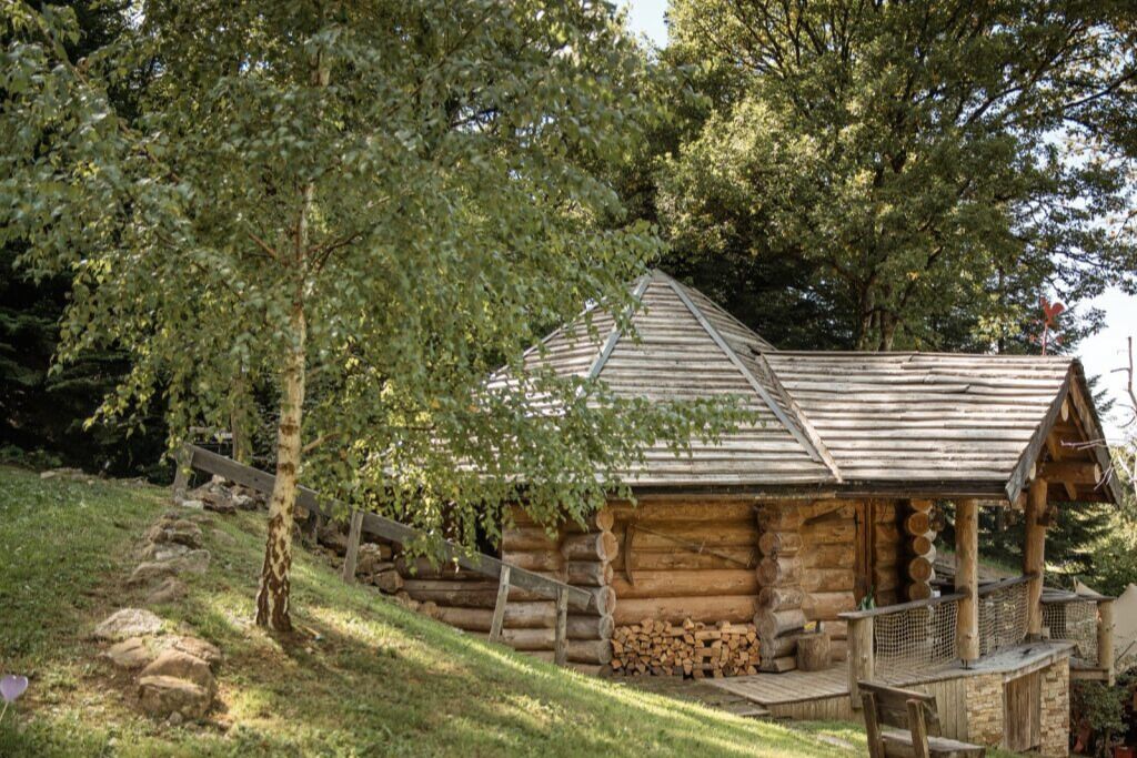 Une cabane rustique en rondins de bois, dotée d’un porche et entourée d’arbres et d’une zone herbeuse.