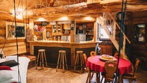 L’intérieur d’un chalet rustique en bois, avec un coin bar, des tabourets en bois, une table ronde recouverte d’une nappe rouge et un escalier.