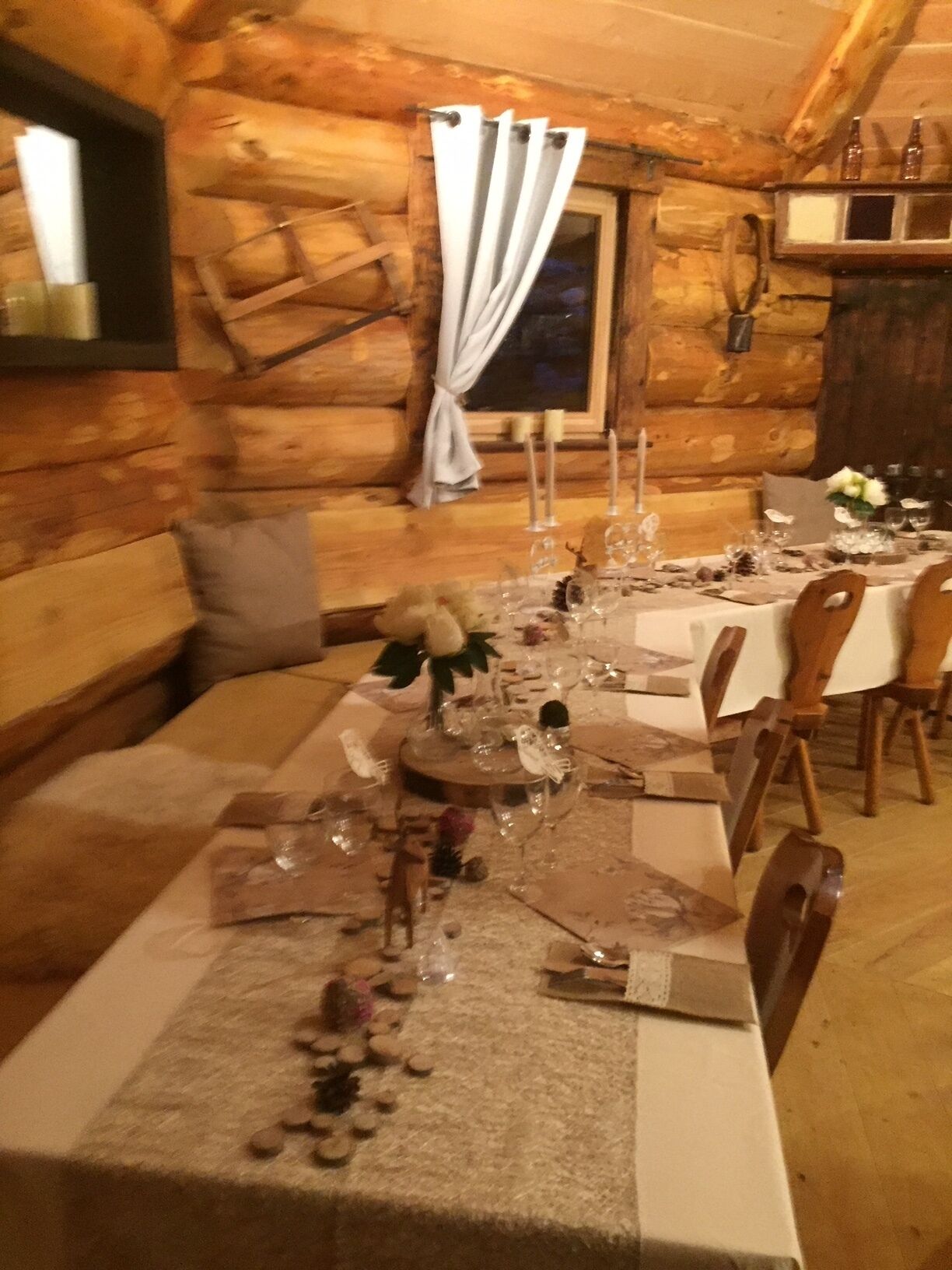 A cozy log cabin dining area with a long table set for a meal, wooden chairs, and a window with curtains.