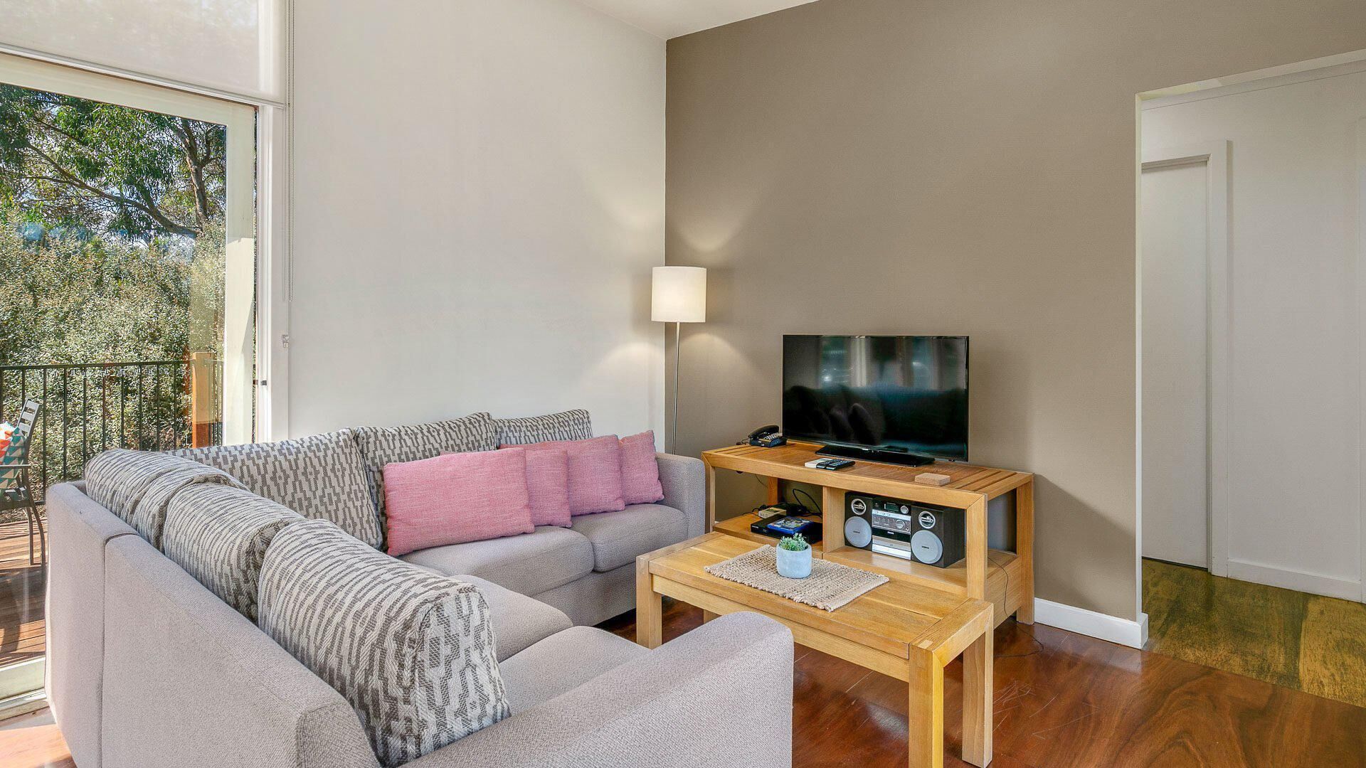Ett modernt vardagsrum med en grå soffa, ett träbord, en tv på ett träställ och en balkong med utsikt över träd.