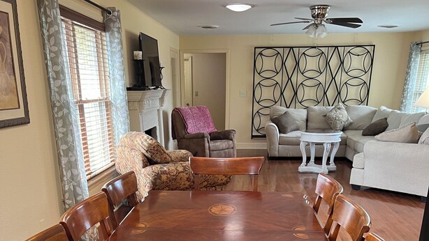 A dining room with a large wooden table surrounded by chairs, a fireplace, and a living area with sofas and a ceiling fan. - Close to a Vibrant and fun DownTown Rose District. 15 min from Tulsa. (Broken Arrow)