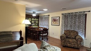A living room with a sofa, patterned cushions, a floral armchair, a dining table with chairs, and a ceiling fan. - Close to a Vibrant and fun DownTown Rose District. 15 min from Tulsa. (Broken Arrow)