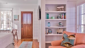 A room with a wooden door, a bookshelf with decorative items, a pink sofa with a fish-patterned pillow, and a patterned rug. - Sweetwater - Pet Friendly in River Dunes Community (Oriental)