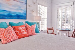 A bedroom with a large bed, multiple pillows, a painting, a window with blinds, a lamp, and a small table with chairs.