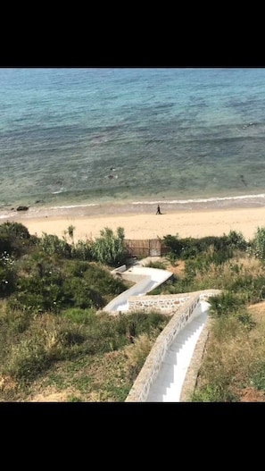 Una playa con agua cristalina, una orilla arenosa y una persona caminando. Hay escaleras que bajan a la playa.