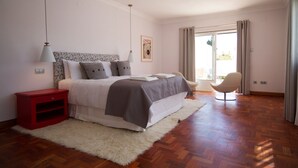 Ein Schlafzimmer mit einem Bett, einem roten Nachttisch, einem weißen Teppich, einem Stuhl und einem großen Fenster mit Vorhängen.