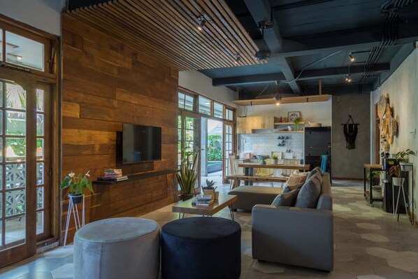 A modern living room with a wooden wall, a flat-screen TV, and a dining area with a kitchen in the background.