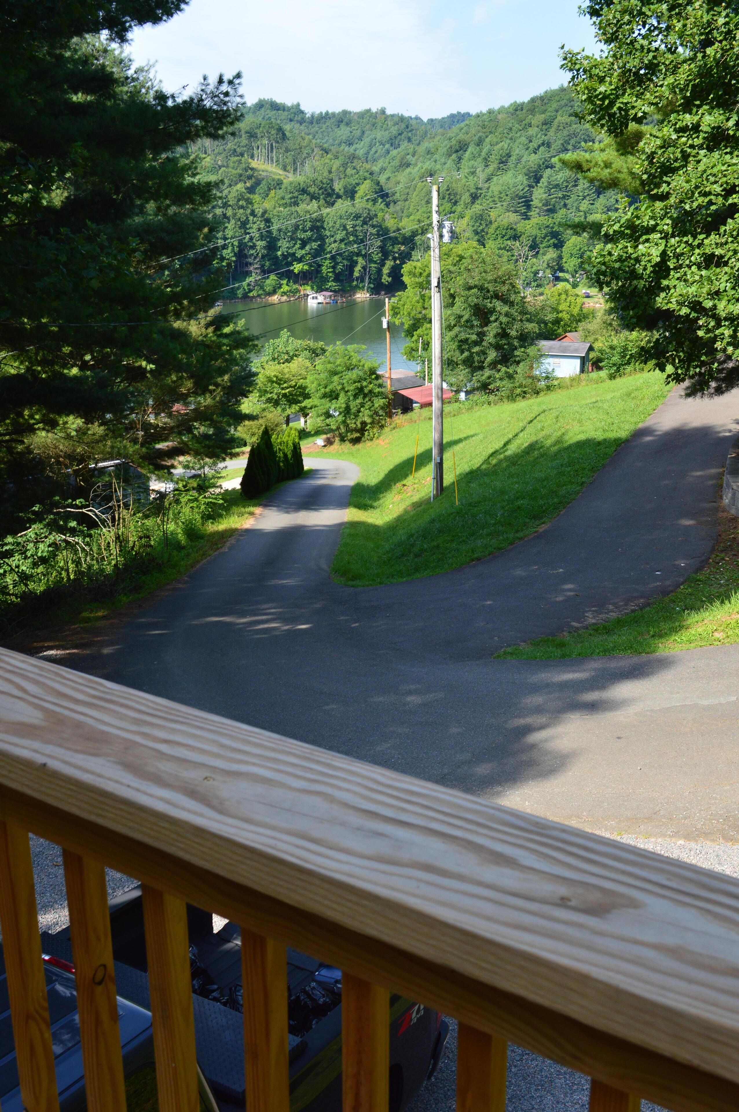 A view from a wooden deck overlooking a winding road, a lake, and a forested hillside.