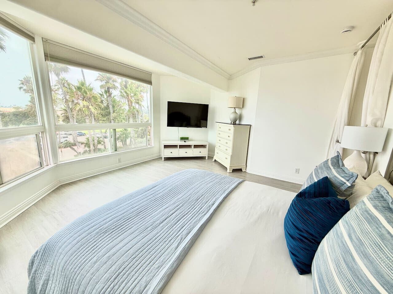 Ein Schlafzimmer mit einem großen Bett, einem Fernseher, einem Schminktisch und einem Fenster mit Blick auf Palmen.