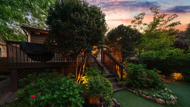 A wooden deck with a staircase leading to a covered area, surrounded by lush greenery and flowering plants. - Classic VAHi Bungalow (Atlanta)