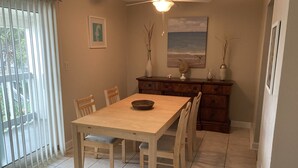 Un comedor con una mesa y sillas de madera, un ventilador de techo, vista al océano a través de la ventana, y un mueble de madera con objetos decorativos.
