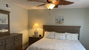 Un dormitorio con cama, mesita de noche con lámpara, cómoda, ventilador de techo y dos cuadros en la pared.