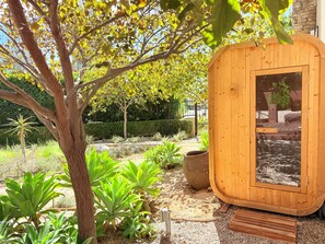 Una sauna de fusta amb porta de vidre, envoltada d'un jardí amb plantes variades i un arbre.