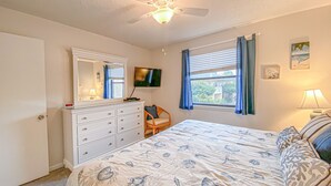 Une chambre à coucher comprenant un lit, une commode, un ventilateur de plafond, une télévision, une fenêtre avec des rideaux et une lampe.
