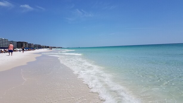 Une plage à l’eau turquoise et limpide, un rivage sablonneux et une jetée visible au loin.