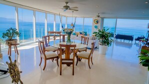 A spacious dining area with a round table and chairs, large windows offering ocean views, and potted plants.