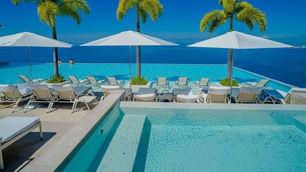 A poolside area with lounge chairs and umbrellas, palm trees, and a clear view of the ocean.