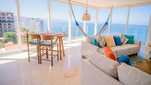 A spacious living room with a large window overlooking the ocean, a white sofa, a wooden table, and a hammock.