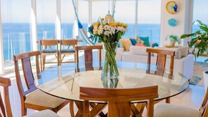 A round glass table with wooden chairs, a vase of flowers, and a view of the ocean through large windows.