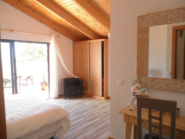 Ein Schlafzimmer mit einem Bett, einem Schreibtisch und einem Stuhl. Eine Schiebetür führt zu einem Balkon mit Sitzgelegenheiten und Topfpflanzen.