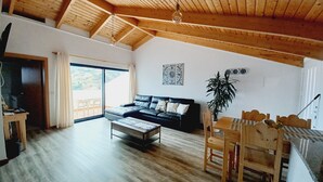 A spacious living room with a high wooden ceiling, a black leather sofa, a wooden dining table with chairs, and a large window offering a view of the outdoors. - Villa Luz II - between Serra and the Sea - Free Wifi (Santa Cruz)
