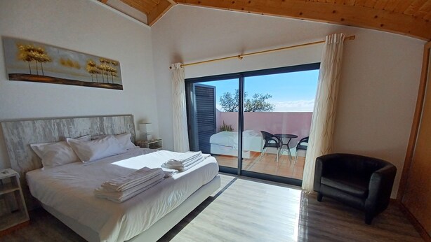 A bedroom with a large bed, a nightstand, a chair, and a view of the outside through a sliding glass door. - Villa Luz II - between Serra and the Sea - Free Wifi (Santa Cruz)