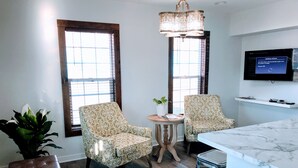 A modern living room with two patterned armchairs, a wooden bench, a small round table with a plant, and a marble kitchen island.