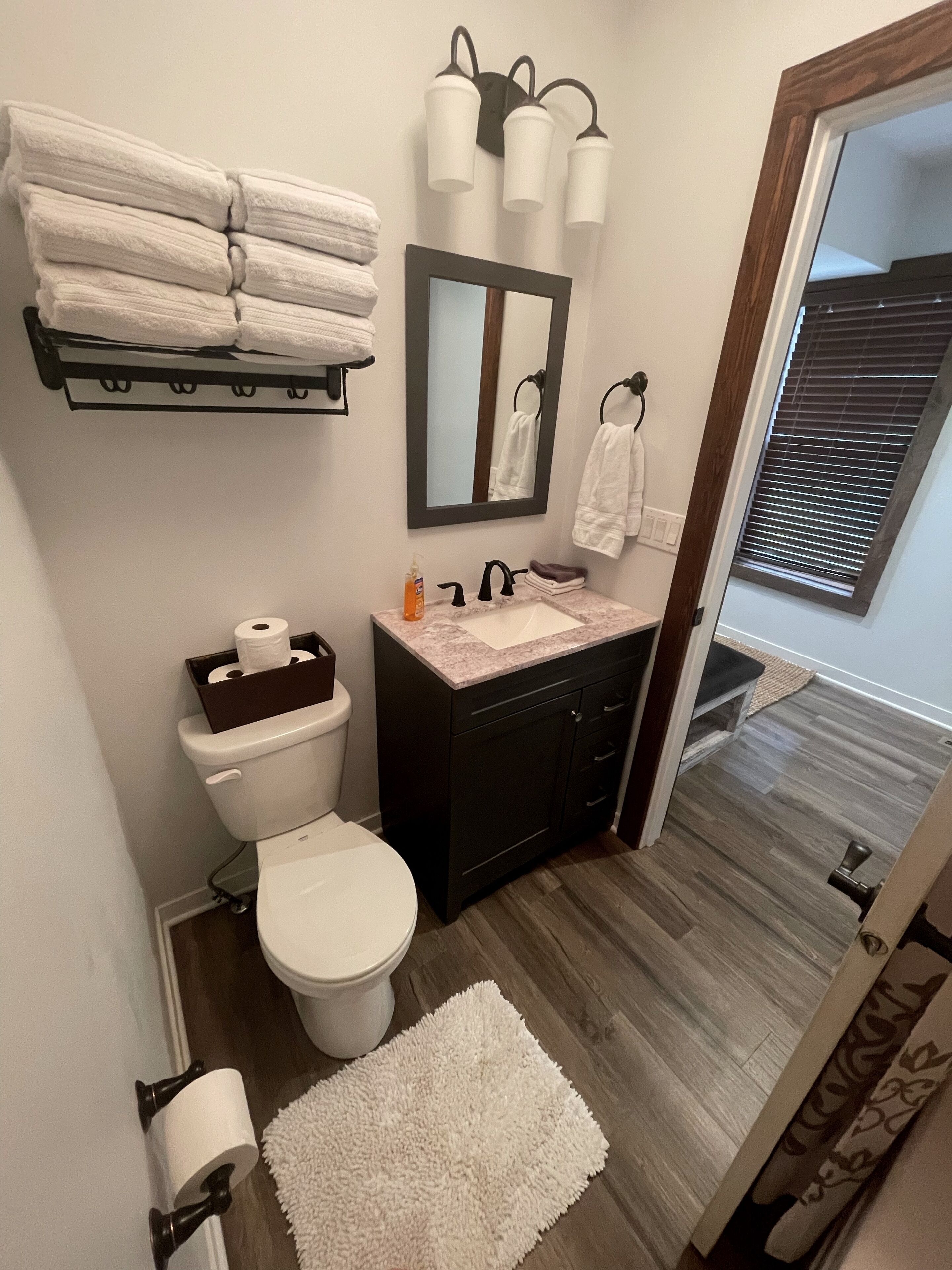 Une salle de bain avec des toilettes, un lavabo et un miroir. Des serviettes sont pliées et empilées sur un porte-serviettes, et un tapis blanc est posé sur le sol.